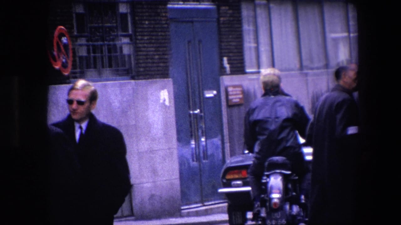1967 Amsterdam street close view with a man in sunglasses, a motorcycle rider seen from behind, pedestrians in coats, a doorway, and a parked dark car.