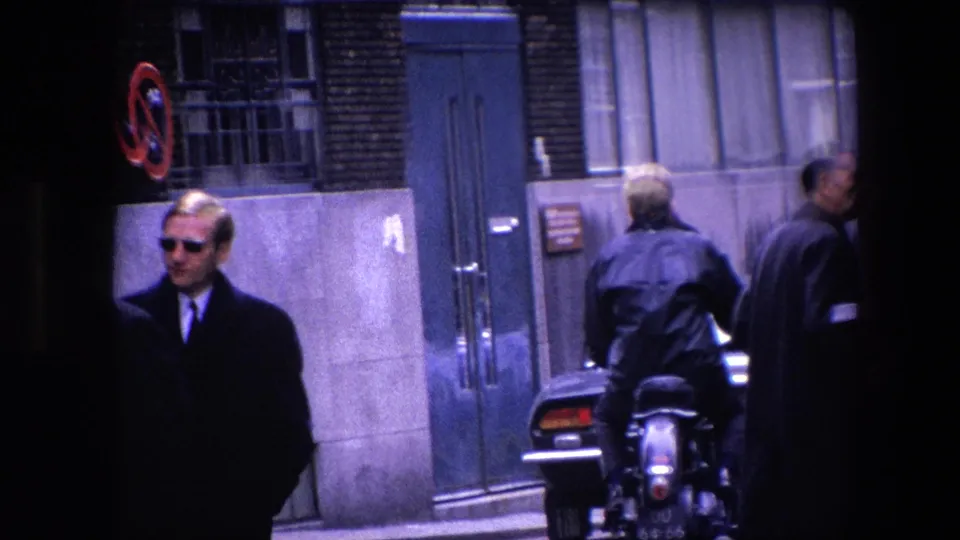 1967 Amsterdam street close view with a man in sunglasses, a motorcycle rider seen from behind, pedestrians in coats, a doorway, and a parked dark car.