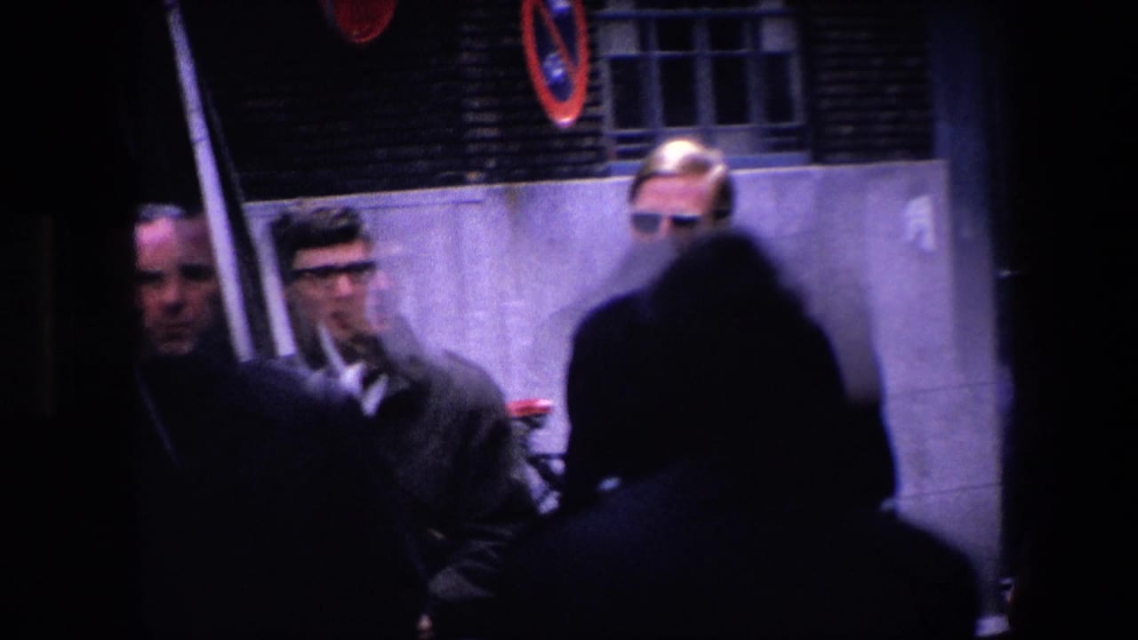 1967 Amsterdam street frame with men in coats near a bicycle, one figure partly blocking the view, and a red no-entry sign on the building behind them.