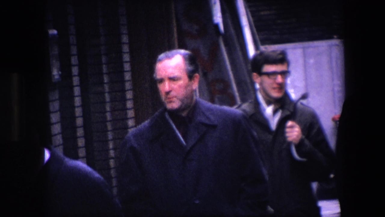 Men in dark coats walk along a narrow city sidewalk with brick buildings behind them.