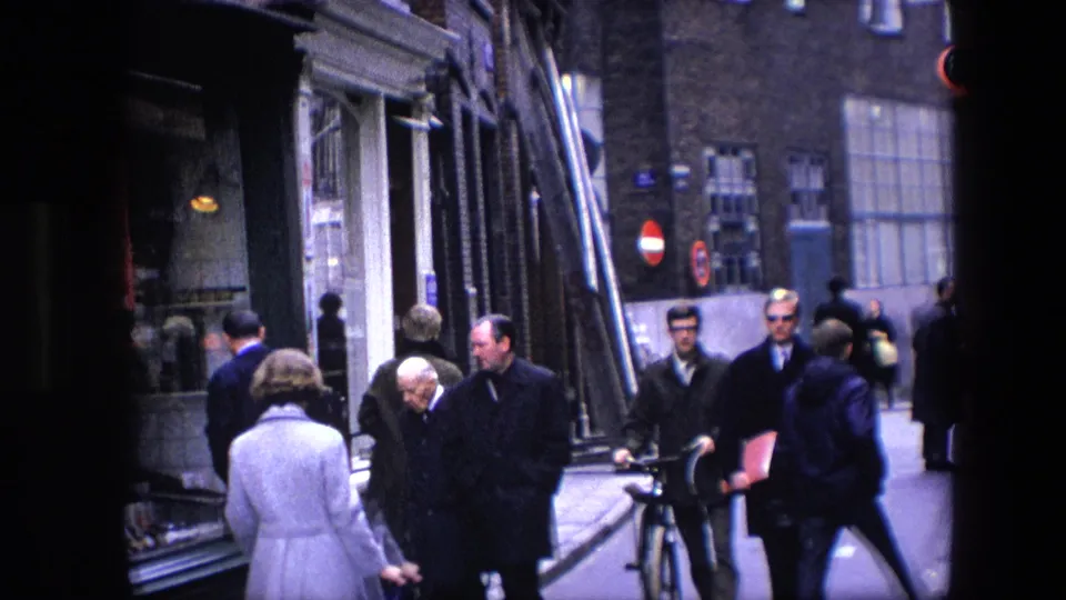 A wider handheld frame captures pedestrians moving past storefront windows, a bicycle, and street signs in footage identified as Amsterdam in 1967.