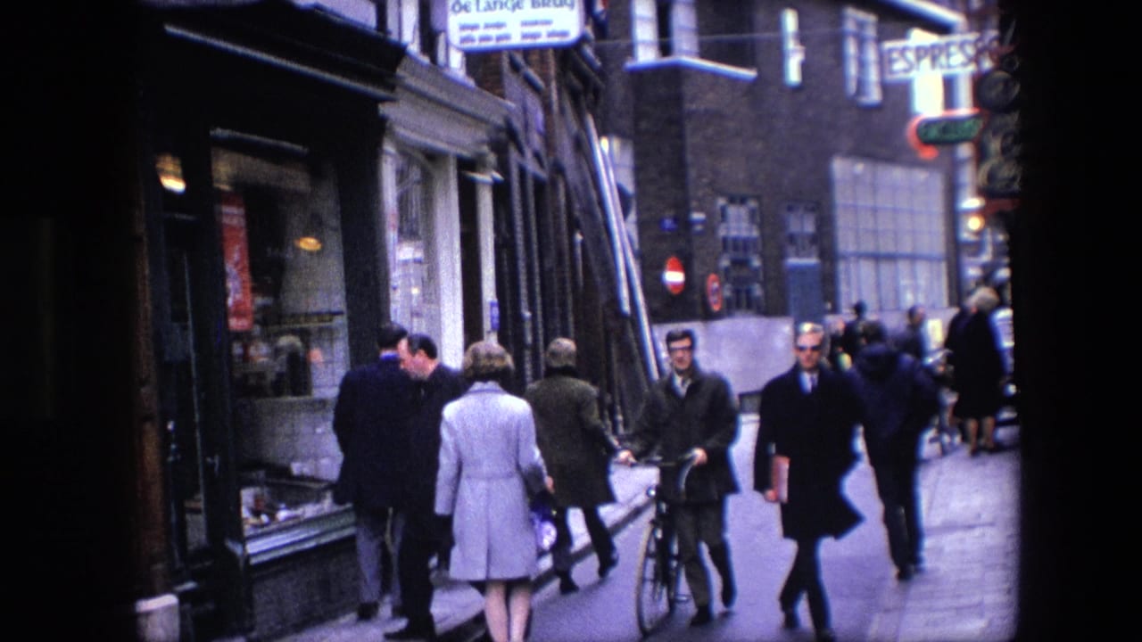 1967 Amsterdam street scene with pedestrians in coats passing shopfronts while a man with a bicycle pauses near the center of the frame.