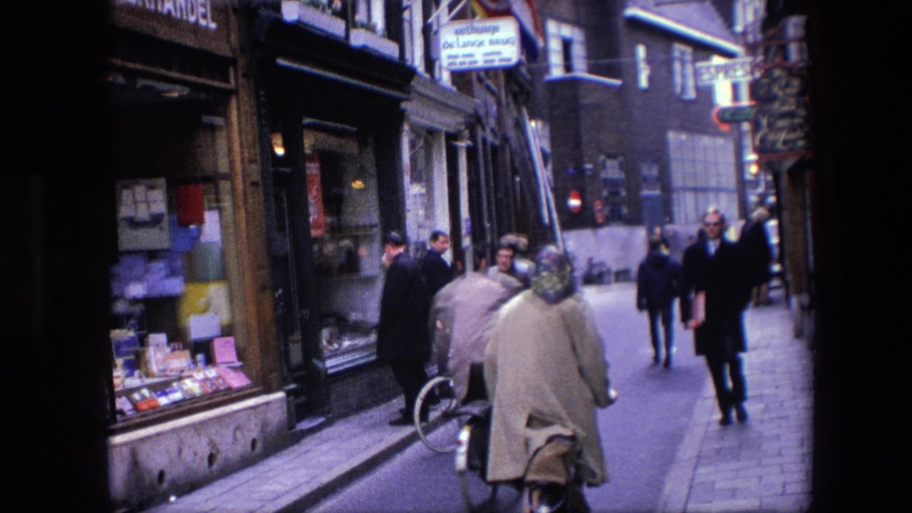 Cyclists and pedestrians pass along a narrow Amsterdam commercial street, with shop windows and overhead signage visible in the 1967 home-movie frame.