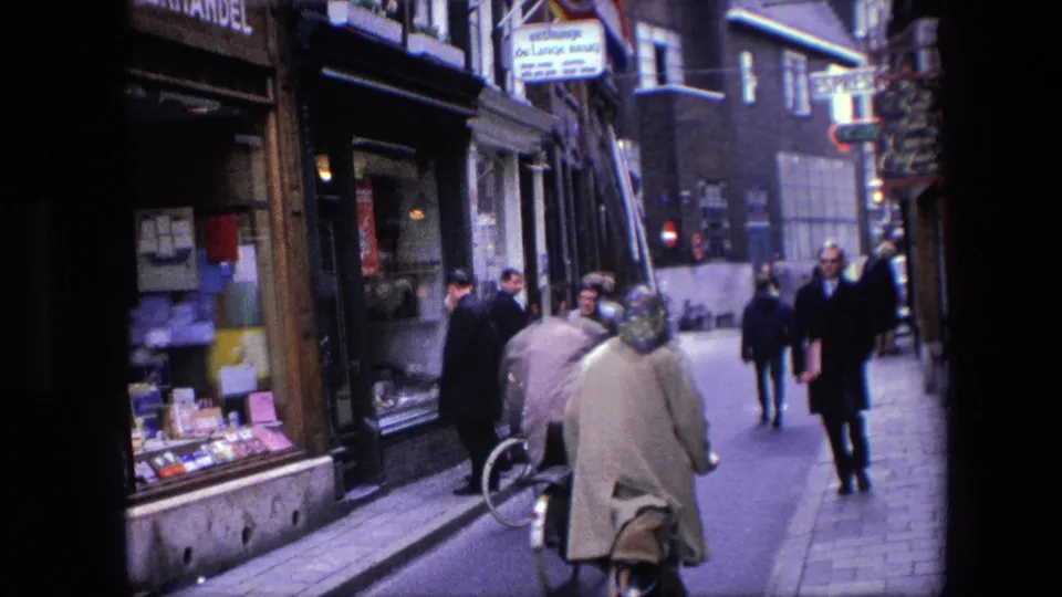 Cyclists and pedestrians pass along a narrow Amsterdam commercial street, with shop windows and overhead signage visible in the 1967 home-movie frame.