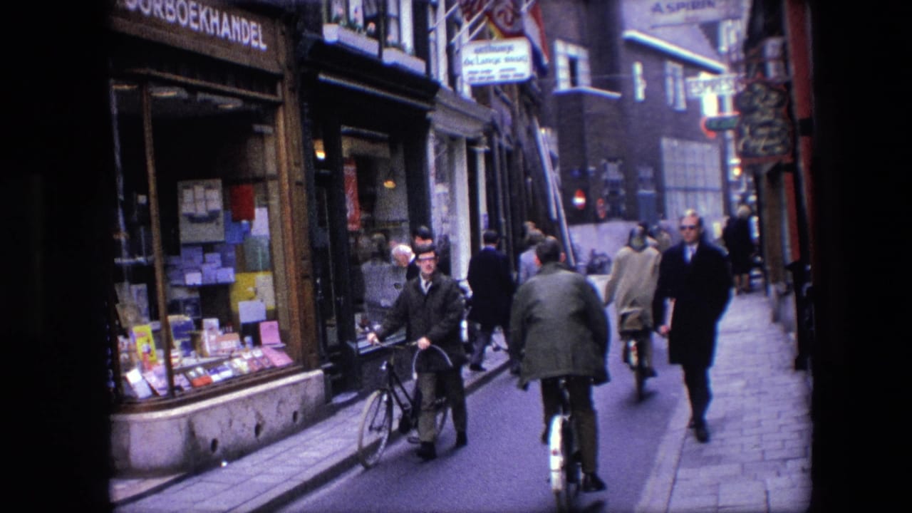 A man walking a bicycle passes a visible bookshop window on a 1967 Amsterdam street as another cyclist and several pedestrians move in the background.