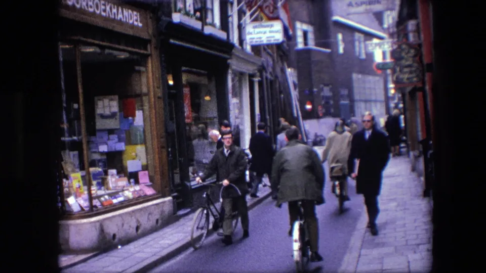 A man walking a bicycle passes a visible bookshop window on a 1967 Amsterdam street as another cyclist and several pedestrians move in the background.