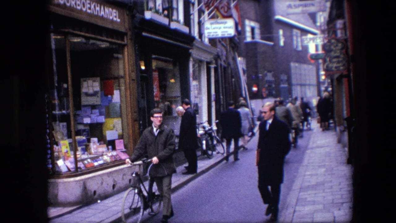 A man with a bicycle occupies the foreground of a 1967 Amsterdam street frame while pedestrians continue past shopfronts and signs in the background.