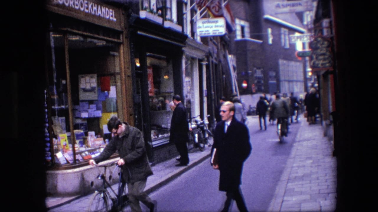 1967 Amsterdam street view with a cyclist near the foreground, pedestrians in coats, and Dutch-language bookshop signage along the left storefront.