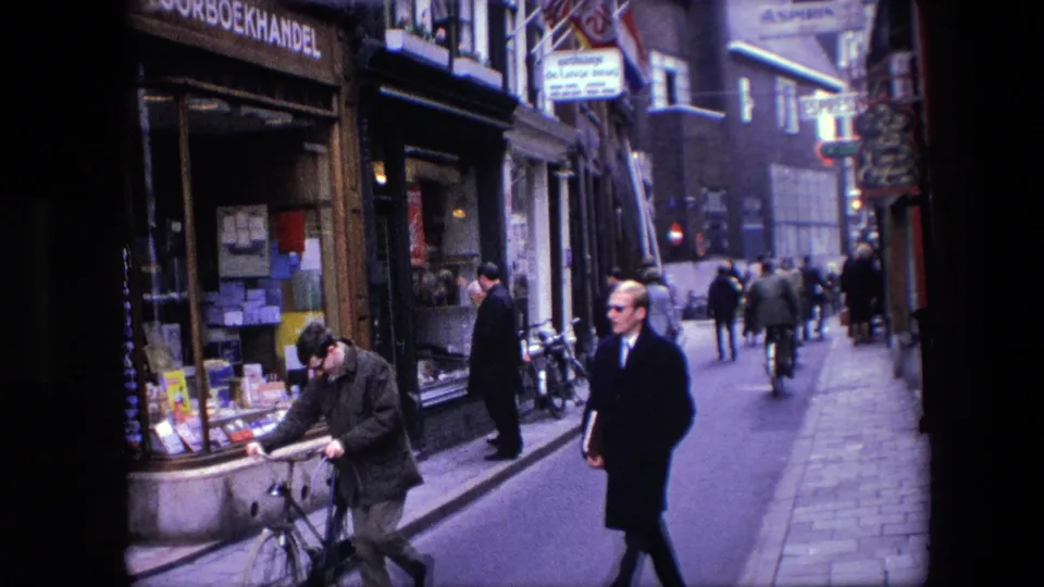 1967 Amsterdam street view with a cyclist near the foreground, pedestrians in coats, and Dutch-language bookshop signage along the left storefront.