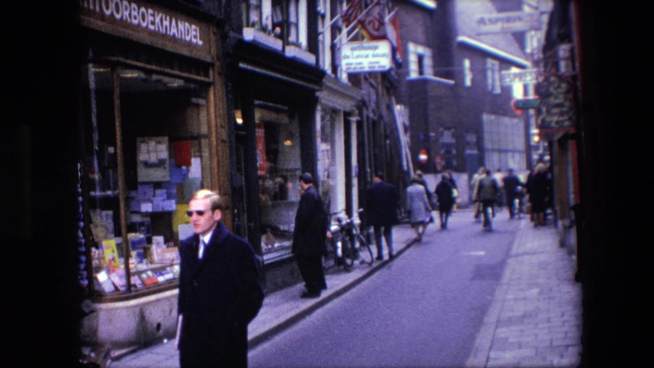 A man in a dark coat and sunglasses walks toward the camera on a narrow Amsterdam street with bicycles, pedestrians, and shopfront signage visible behind him.