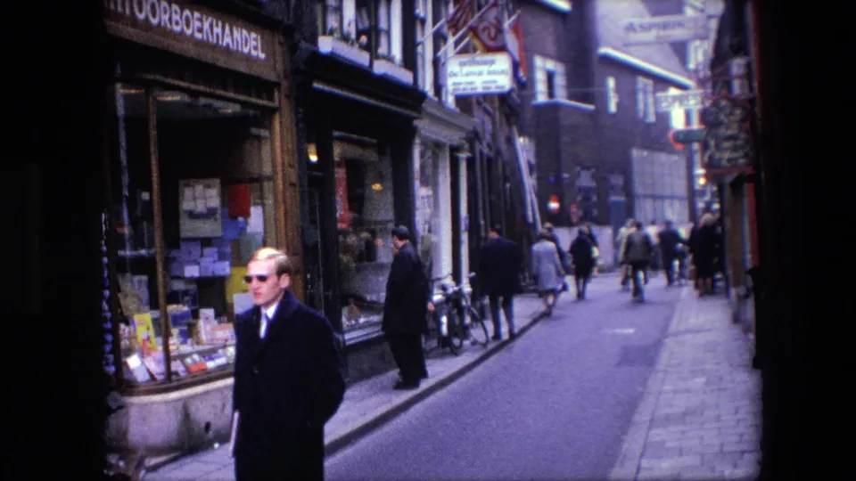 A man in a dark coat and sunglasses walks toward the camera on a narrow Amsterdam street with bicycles, pedestrians, and shopfront signage visible behind him.