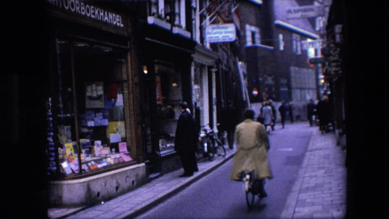 A cyclist wearing a light coat rides away along a narrow Amsterdam street, with pedestrians, bicycles, and a bookshop frontage visible in the background.