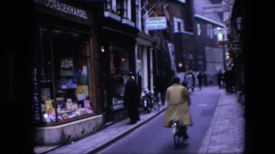 A cyclist wearing a light coat rides away along a narrow Amsterdam street, with pedestrians, bicycles, and a bookshop frontage visible in the background.