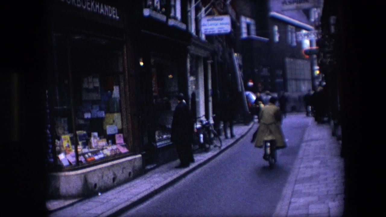 1967 Amsterdam street scene with a cyclist in a light coat riding down a narrow street past shop windows and pedestrians.