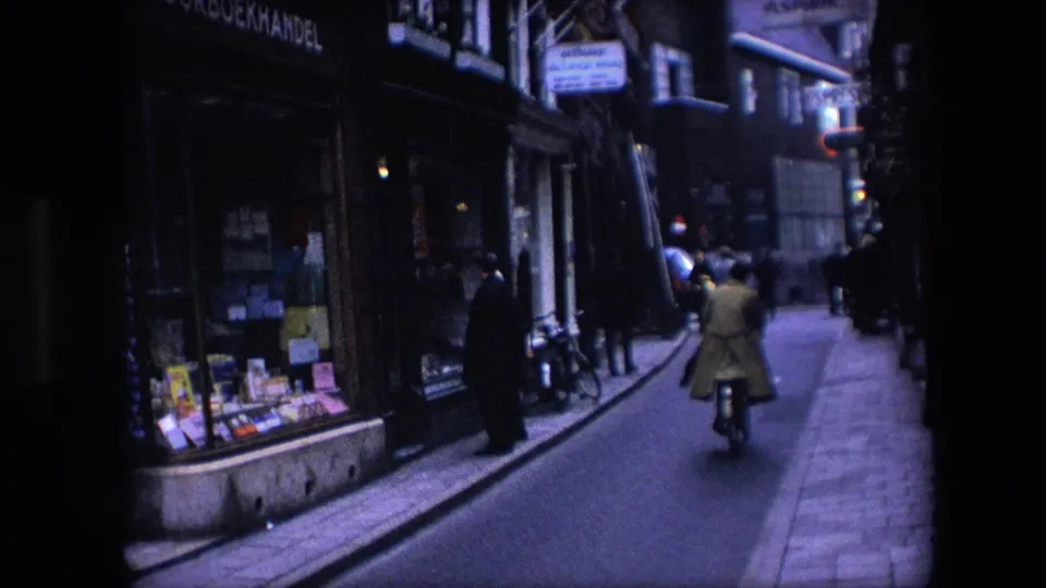 1967 Amsterdam street scene with a cyclist in a light coat riding down a narrow street past shop windows and pedestrians.