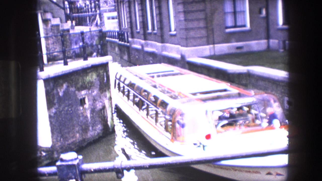 Canal tour boat with passengers moves through a narrow Amsterdam canal or lock area beside masonry buildings and railings.
