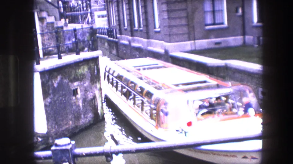 Canal tour boat with passengers moves through a narrow Amsterdam canal or lock area beside masonry buildings and railings.