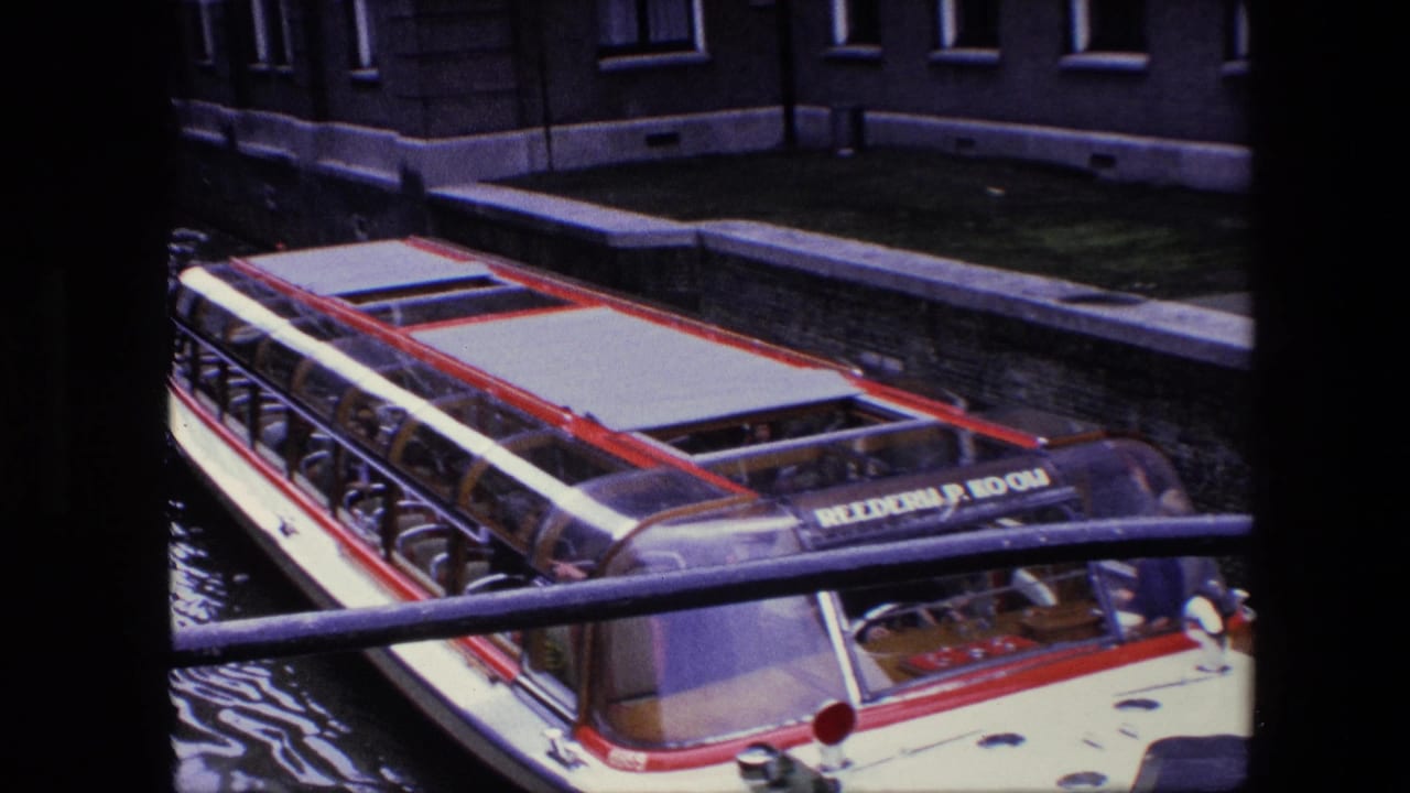 Amsterdam canal boat passes close below a railing, with passengers visible through the glass roof and canal-side buildings behind it.