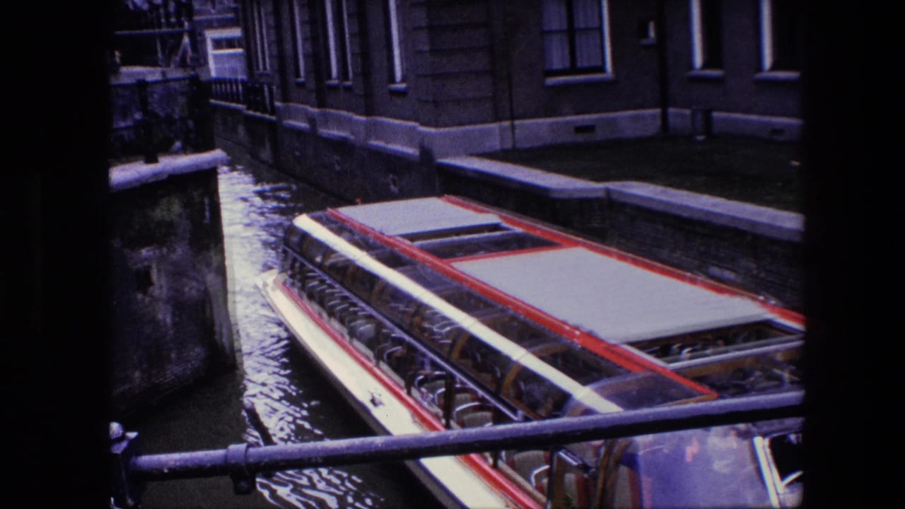 1967 Amsterdam canal scene with a red and white sightseeing boat moving beneath a dark foreground structure, with passengers visible through the glass canopy.