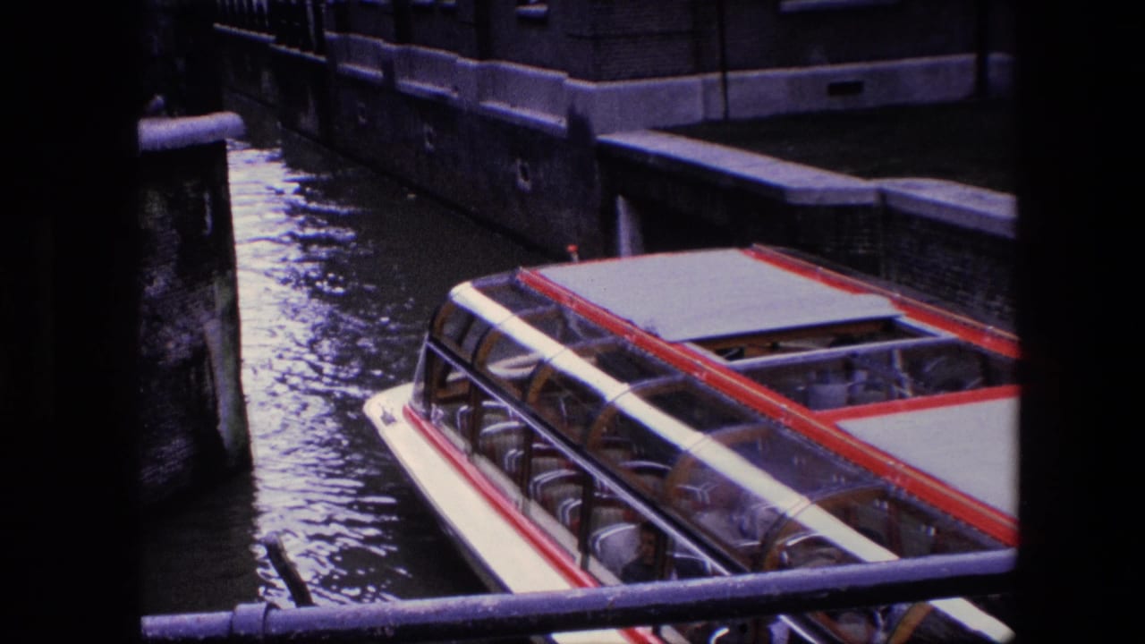 A red and white canal tour boat with passengers under a glass canopy moves through the Amsterdam canal, seen from an elevated home-movie angle.