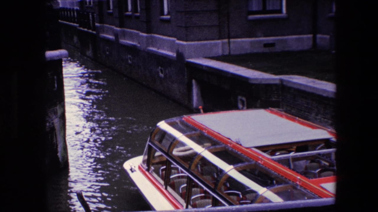 The sightseeing boat moves out of frame along the Amsterdam canal, leaving rippled water, canal walls, and building windows visible behind it.
