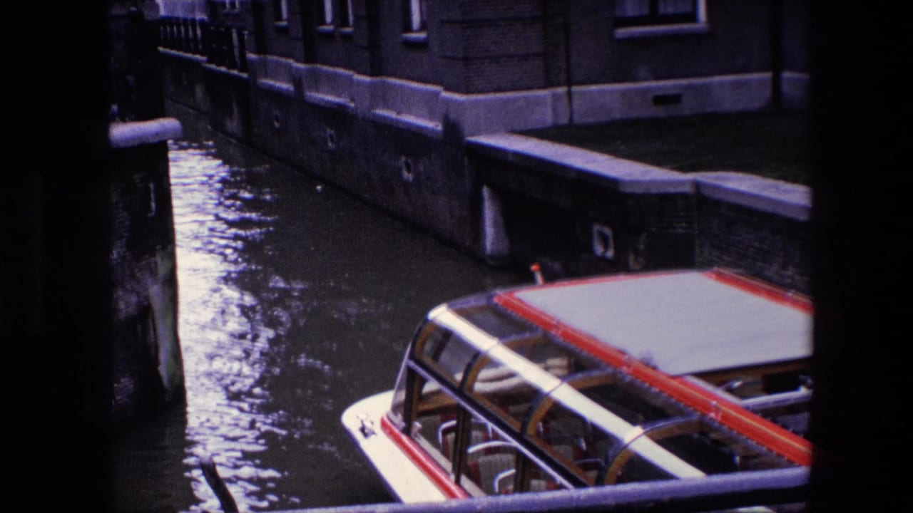 1967 8mm Amsterdam canal view showing the roof of an enclosed passenger boat moving through water beside masonry walls and tall-windowed buildings.