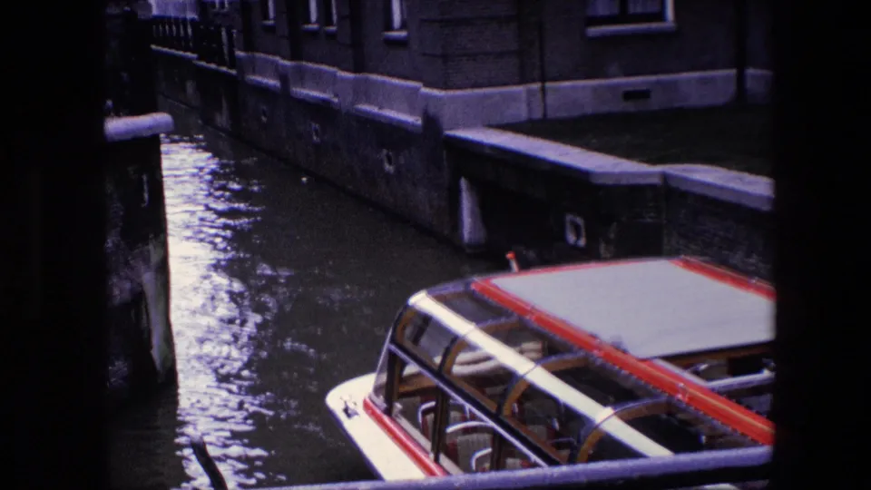 1967 8mm Amsterdam canal view showing the roof of an enclosed passenger boat moving through water beside masonry walls and tall-windowed buildings.