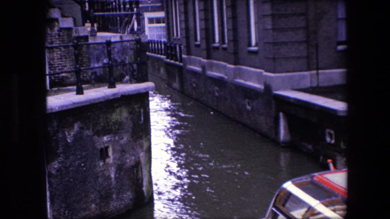 1967 Amsterdam canal scene with an enclosed passenger boat partly visible at the lower right, framed by canal water, brick facades, stone edging, and iron railings.