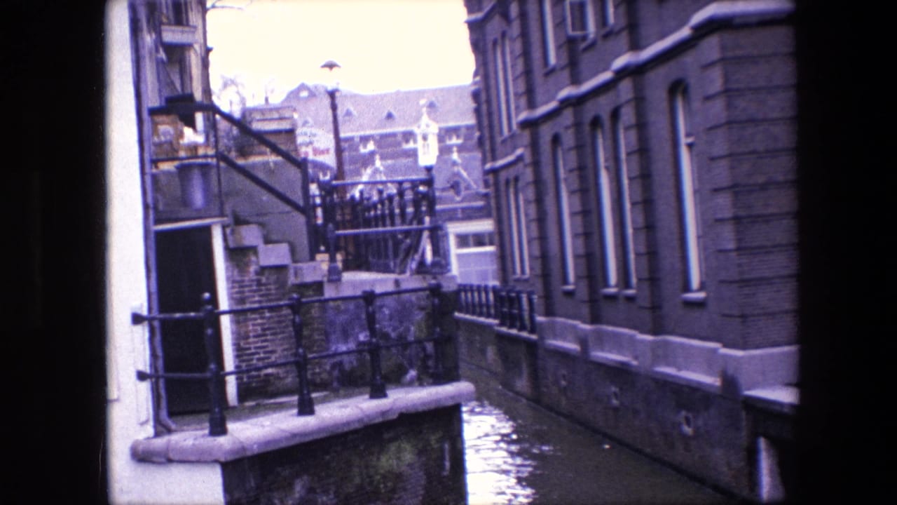 1967 8mm view over an Amsterdam canal with stone steps, black iron railings, masonry buildings, tall windows, and water running between the structures.