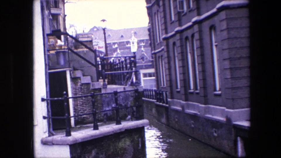 1967 8mm view over an Amsterdam canal with stone steps, black iron railings, masonry buildings, tall windows, and water running between the structures.