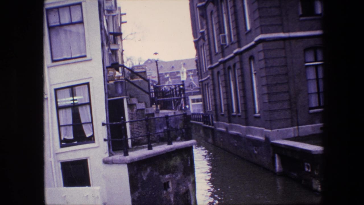 Supplied 1967 Amsterdam footage frame showing a narrow canal, pale building facade, darker masonry building, railings, and still floodwater below.