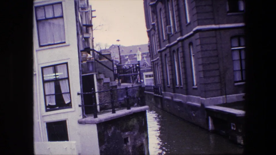 Supplied 1967 Amsterdam footage frame showing a narrow canal, pale building facade, darker masonry building, railings, and still floodwater below.