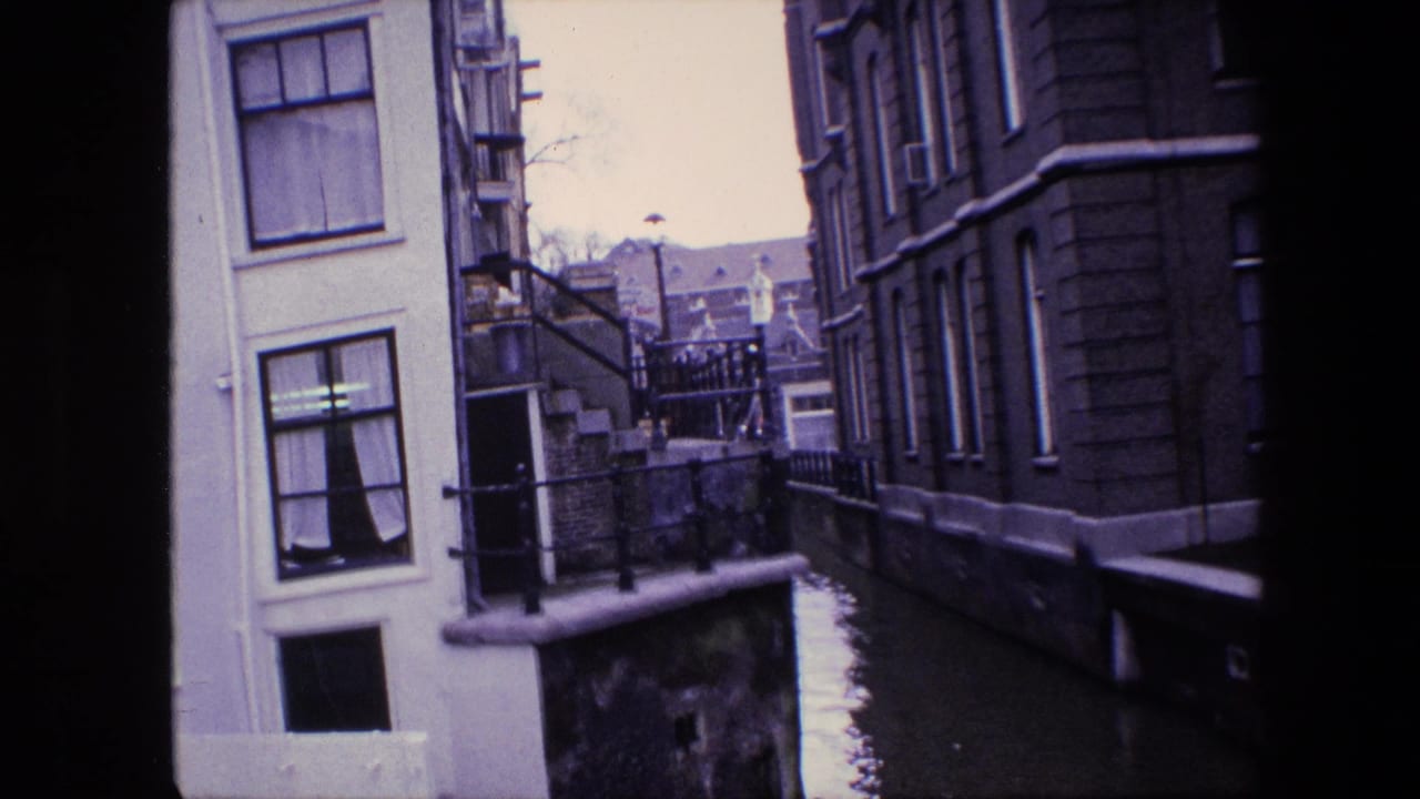 1967 Amsterdam canal view framed between tall buildings, showing curtained windows, railings, canal water, and a small bridge in the distance.