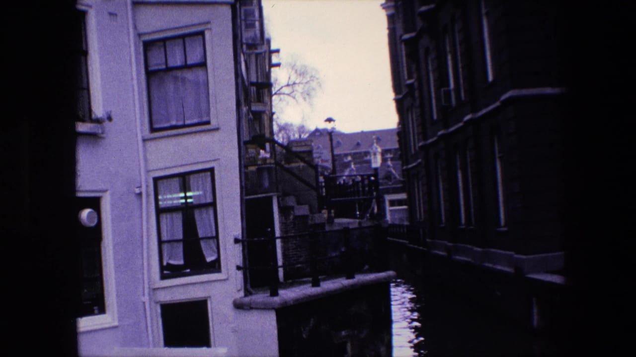 1967 Amsterdam canal-side view with a pale building, dark neighboring facade, railings, water, and distant rooftops seen through a shaded gap.