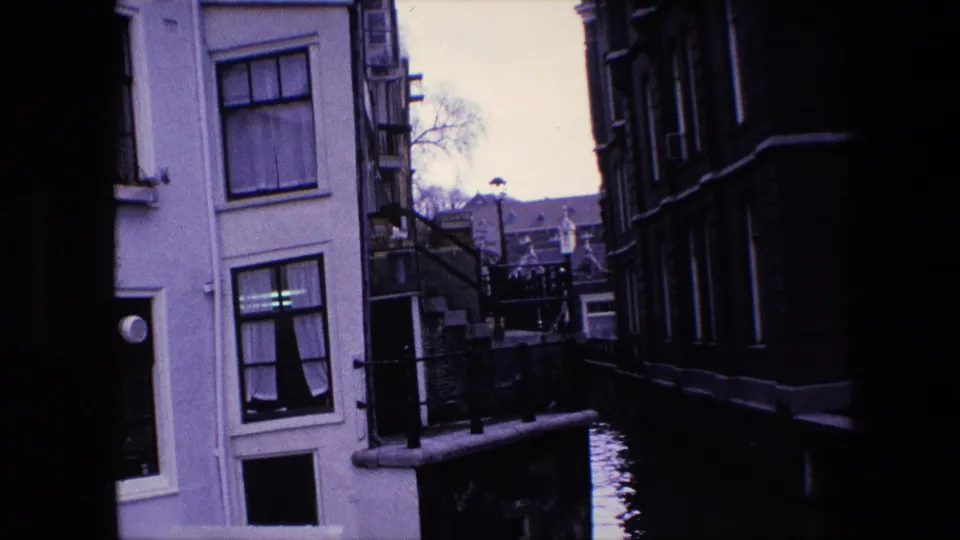 1967 Amsterdam canal-side view with a pale building, dark neighboring facade, railings, water, and distant rooftops seen through a shaded gap.