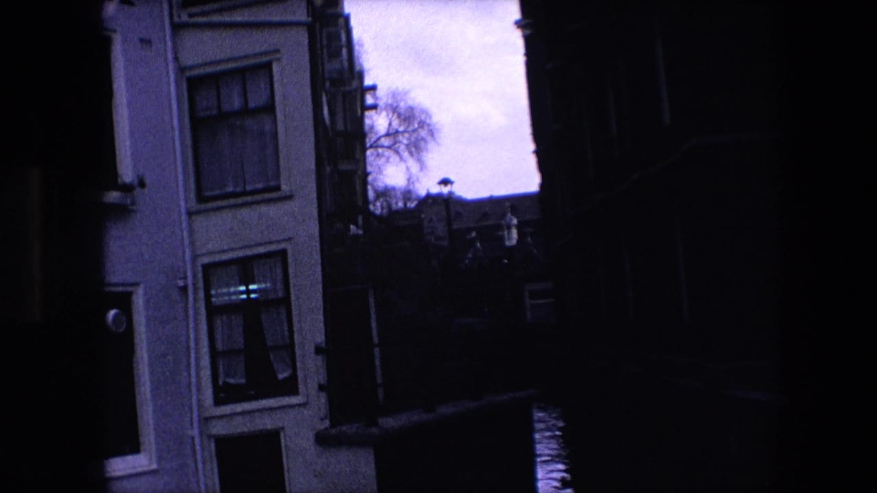 Dim 1967 Amsterdam canal frame showing a pale building with windows, a dark passage, canal water, railing, and distant rooflines under a bright sky.