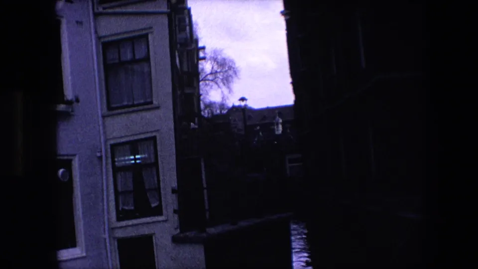 Dim 1967 Amsterdam canal frame showing a pale building with windows, a dark passage, canal water, railing, and distant rooflines under a bright sky.