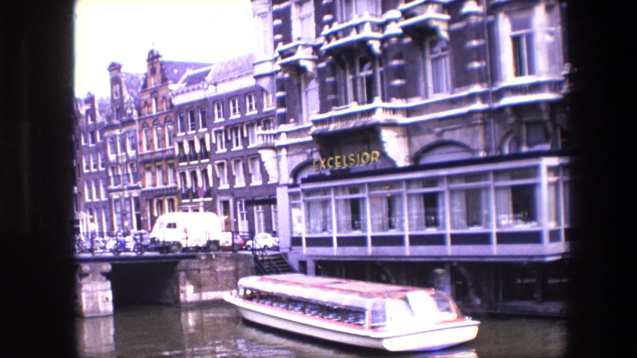 1967 Amsterdam canal scene with a tour boat passing below ornate buildings, parked cars, a white van, bridge railings, and a visible Excelsior hotel sign.