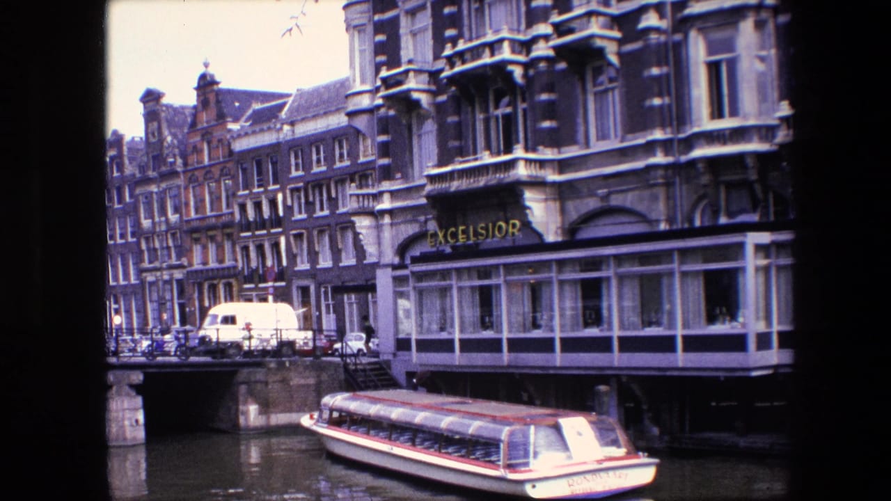 1967 Amsterdam canal view with a white tour boat, stone bridge, bicycles, a white van and the Excelsior hotel frontage visible across the water.