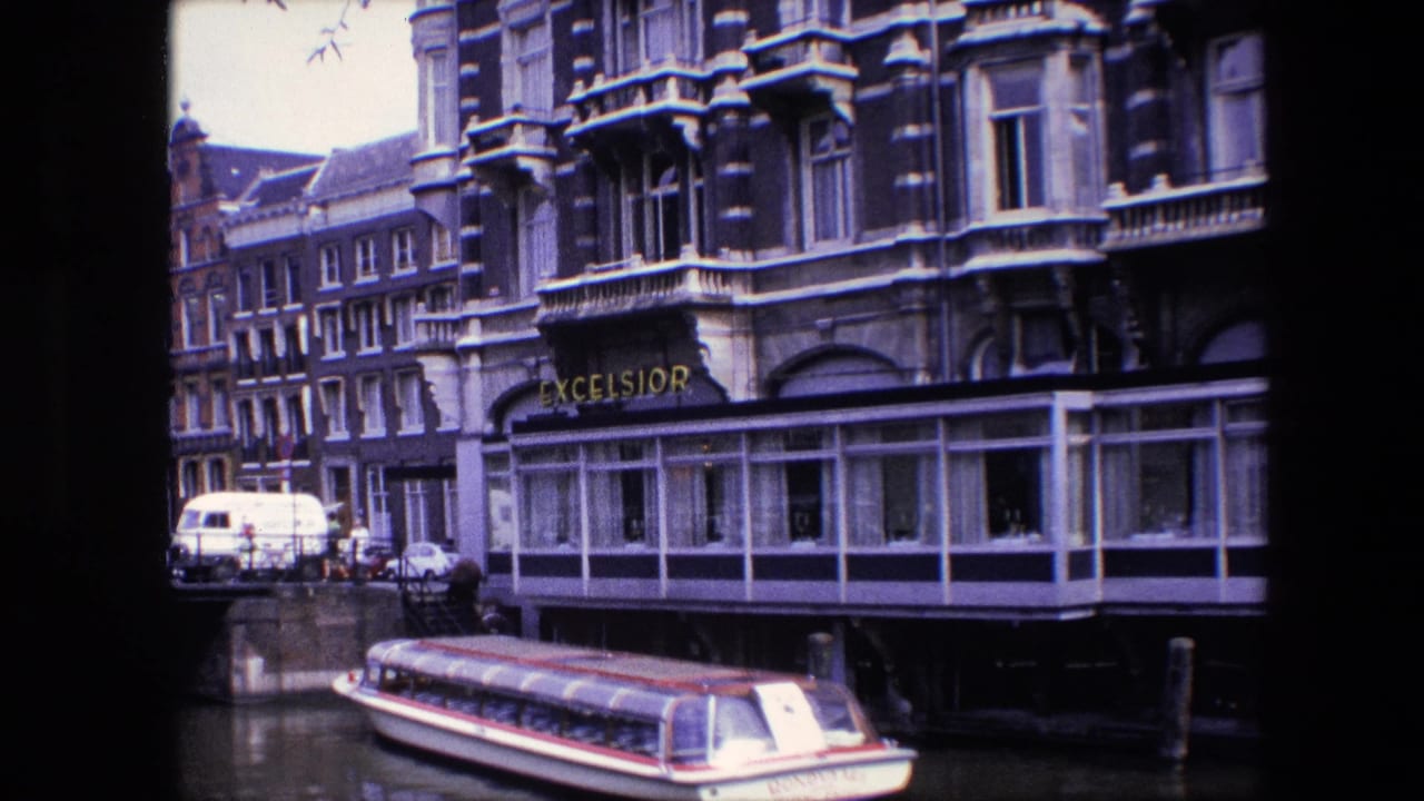 1967 Amsterdam canal footage with the tour boat lower in frame, the Excelsior sign partly visible and dense older buildings lining the canal behind it.