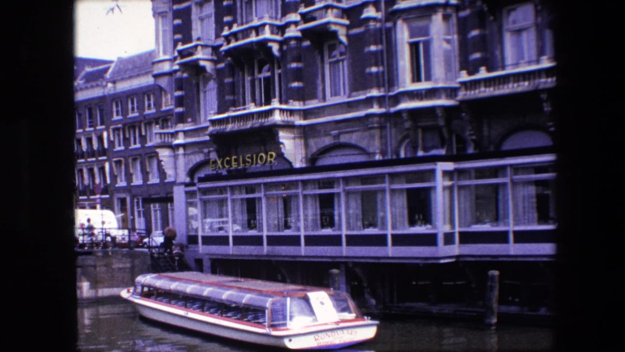 Late pan frame from 1967 Amsterdam showing the tour boat beside the canal wall, Excelsior hotel frontage, bicycles and a white van near the bridge.