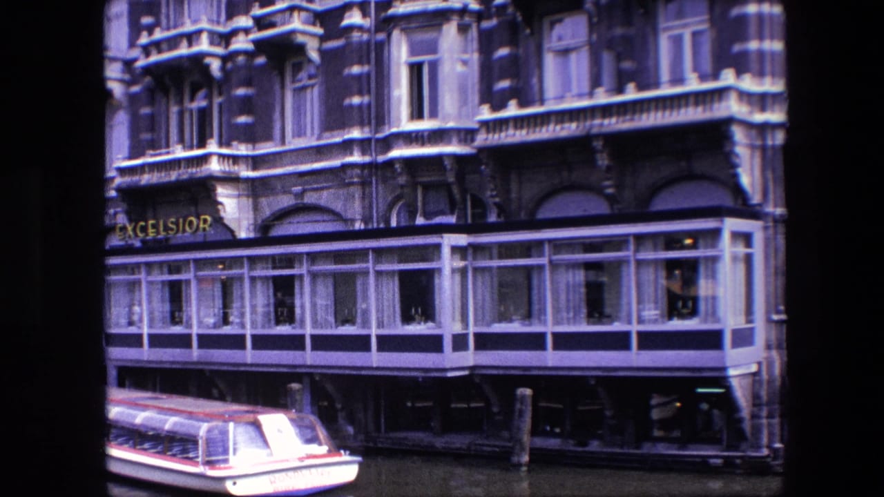 1967 Amsterdam canal footage frame showing the Excelsior hotel sign, ornate upper stories, waterfront windows, and the tail end of a covered tour boat at the bottom of the image.