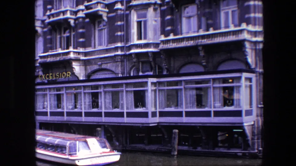 1967 Amsterdam canal footage frame showing the Excelsior hotel sign, ornate upper stories, waterfront windows, and the tail end of a covered tour boat at the bottom of the image.