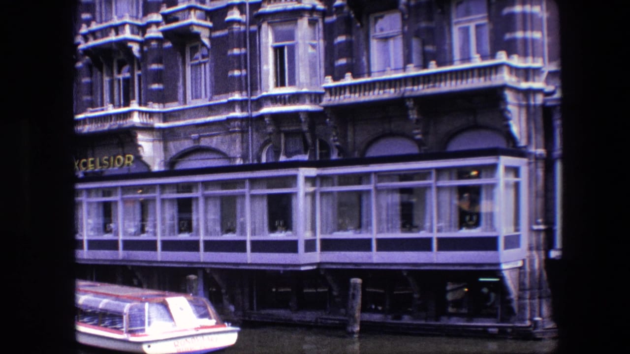 1967 Amsterdam canal view with a small white-and-red boat near mooring posts and a many-windowed glass extension below an ornate building facade.