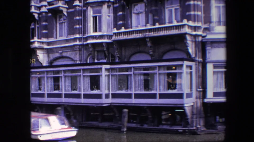 Amsterdam canal-side facade in 1967, showing the glass-enclosed lower level, dark awnings, mooring posts, and the edge of a small boat at left.