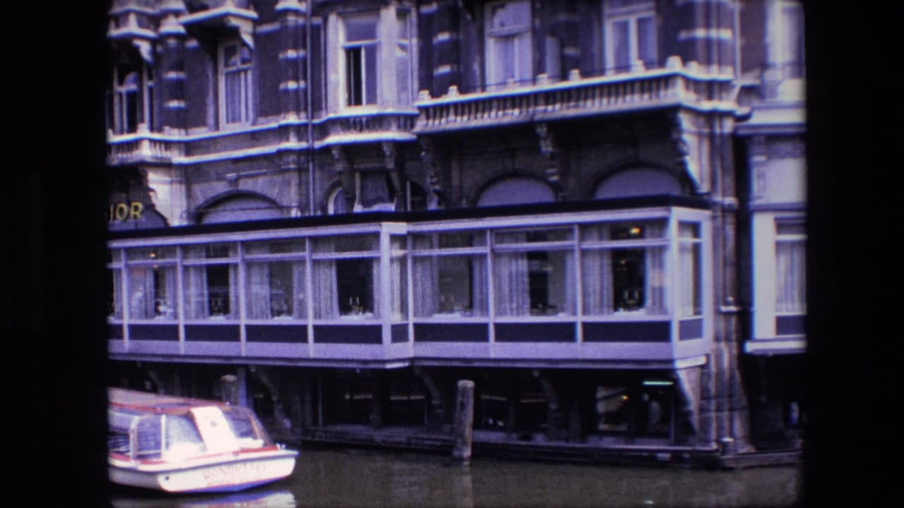 Visitor-shot 8mm view of an Amsterdam canal building, with a small boat partly cropped at lower left and a long glass-fronted section over the water.