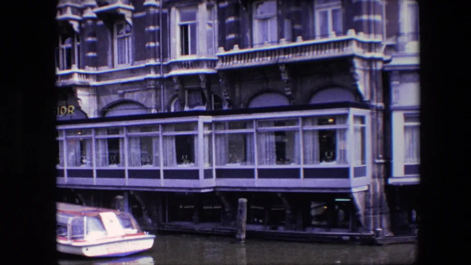Visitor-shot 8mm view of an Amsterdam canal building, with a small boat partly cropped at lower left and a long glass-fronted section over the water.