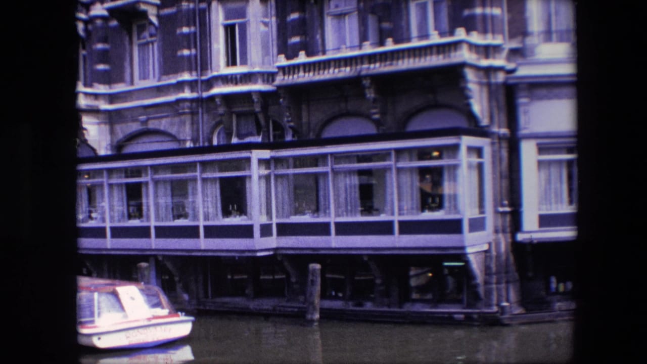 Late frame from the 1967 Amsterdam canal shot, with the glass-fronted building extension centered above calm water and the small boat cropped at the lower left.