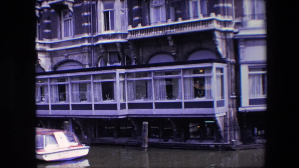 Late frame from the 1967 Amsterdam canal shot, with the glass-fronted building extension centered above calm water and the small boat cropped at the lower left.
