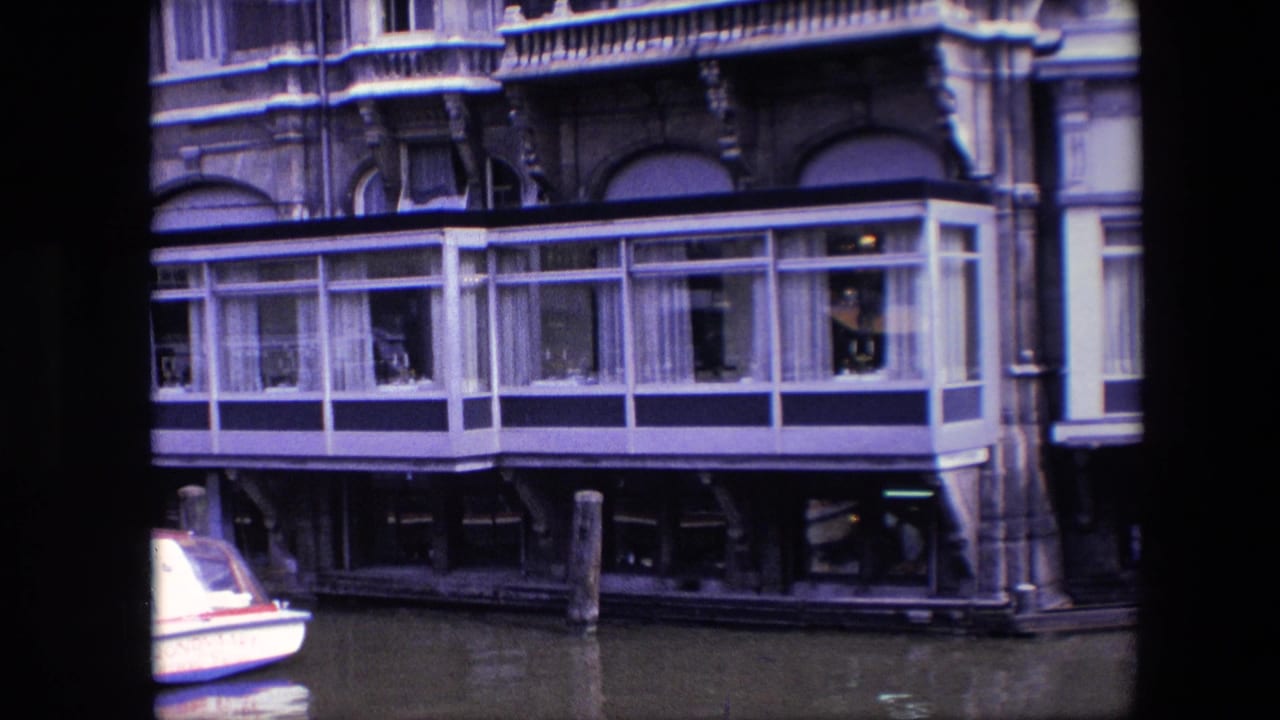 1967 Amsterdam home-movie frame showing a canal houseboat with a glass-enclosed room, white curtains, dark lower supports, mooring posts, and a partial white boat at left.
